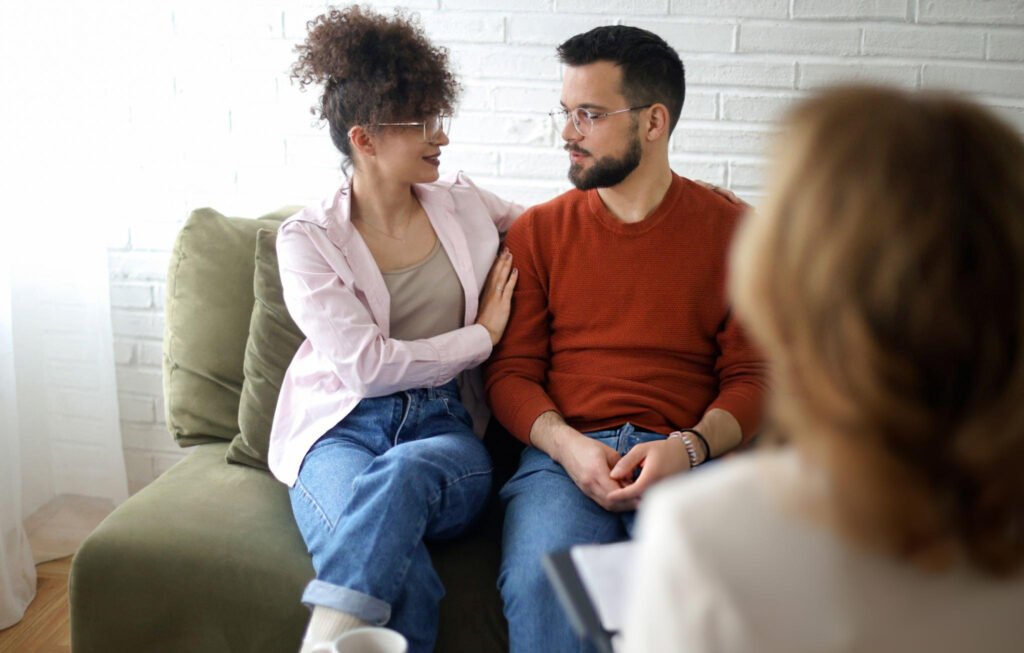 A couple sits together on a couch, exchanging looks of understanding and hope, as a therapist from family counseling services listens attentively in the foreground.