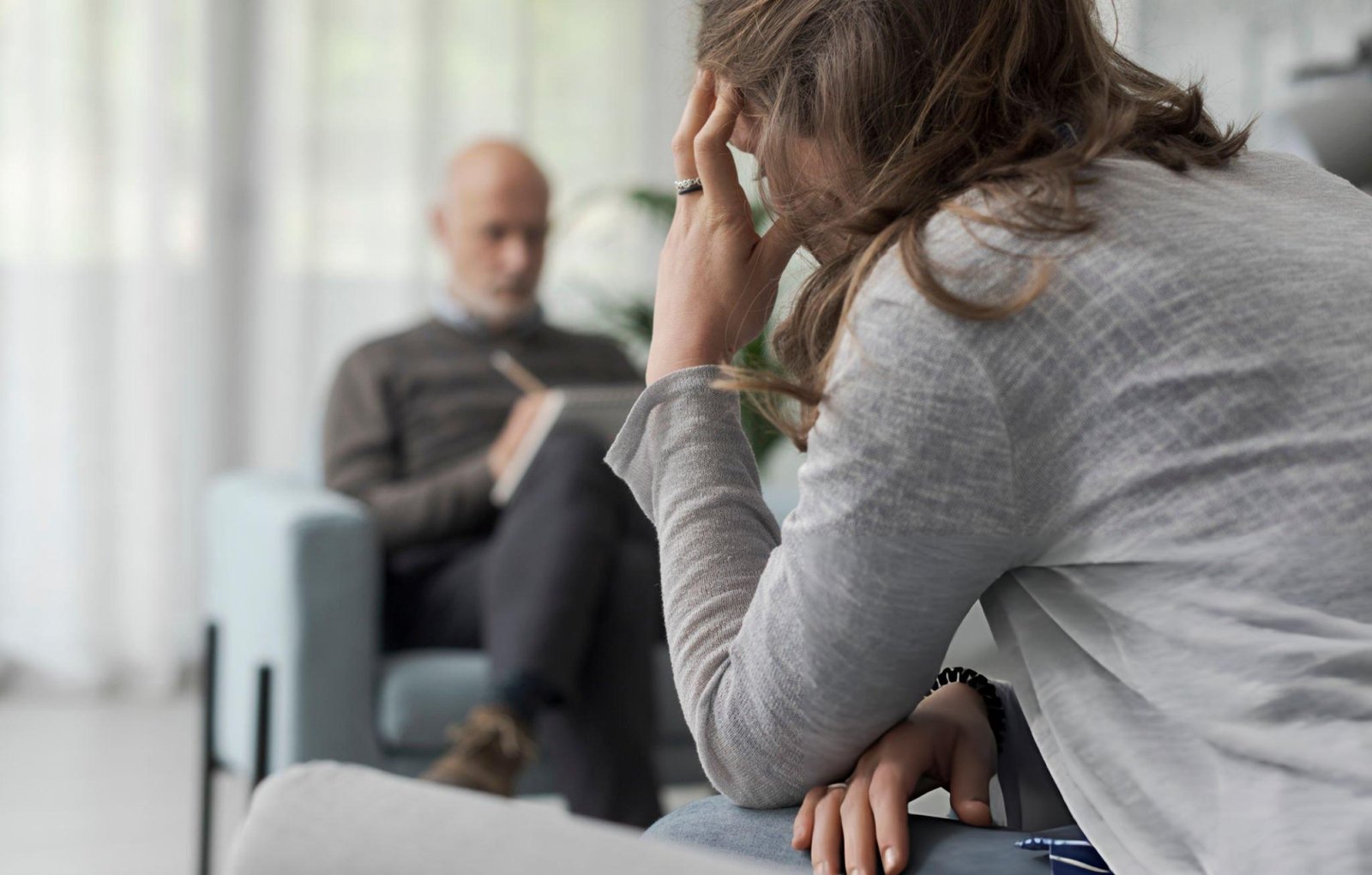 A person is sitting on a couch with a hand on the forehead, appearing to be in a thoughtful or distressed state. In the background, another person is seated on a chair, holding a notepad and pen, suggesting a therapy session. The setting appears to be a professional environment with soft lighting and a calm atmosphere. This image is relevant as it depicts a scene of depression therapy in Chicago.